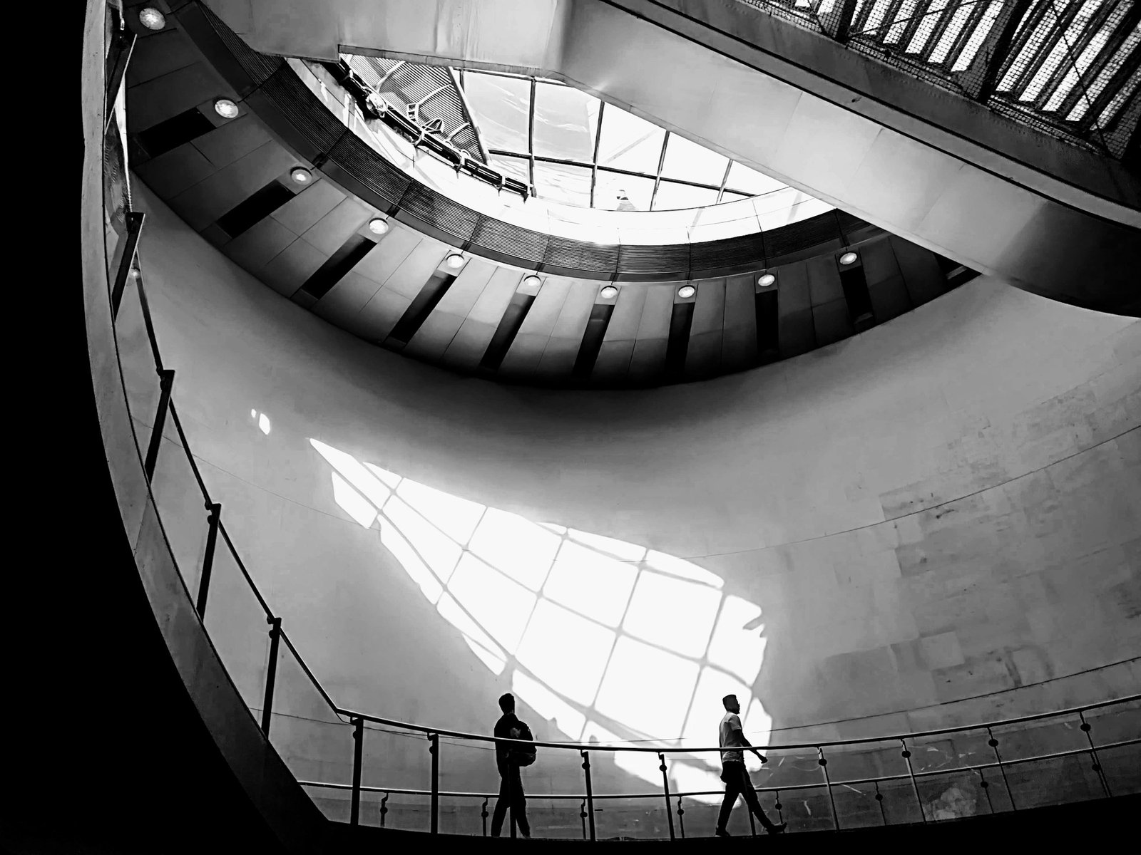 Escalier hélicoïdal en béton sous une verrière, deux silhouettes sur la rampe